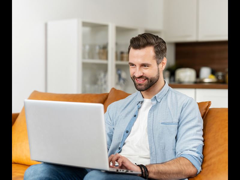 Smiling Young Bearded Hipster Man Sitting On The Couch On Laptop