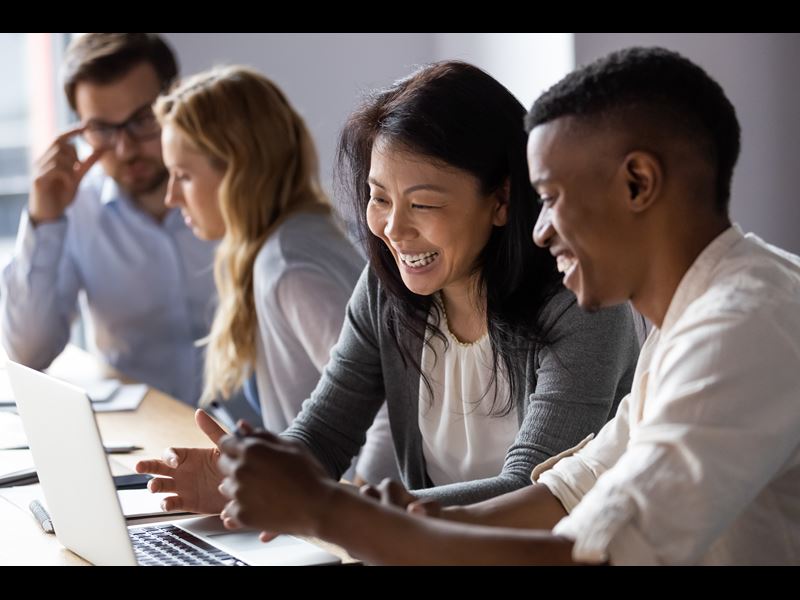 Two employees laughing together while looking at laptop, with two coworkers in the background