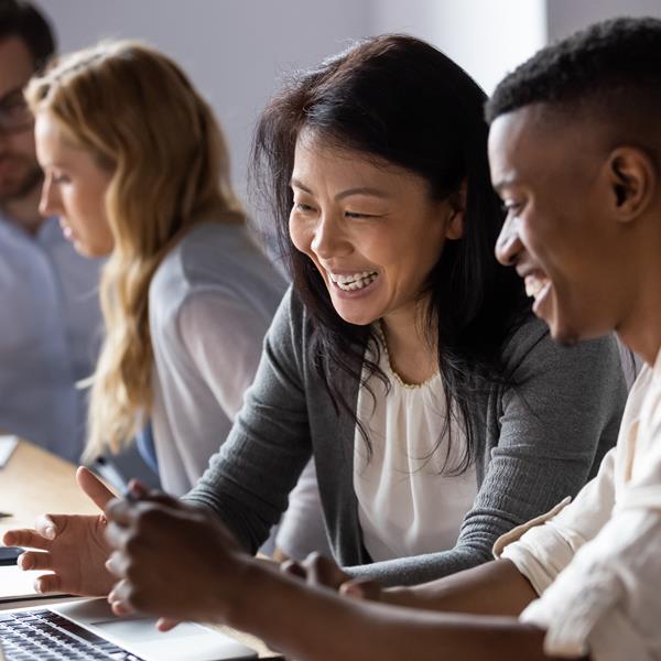 Two employees laughing together while looking at laptop, with two coworkers in the background