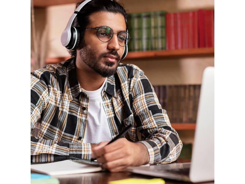 Man wearing glasses and headphones, looking at laptop and taking notes