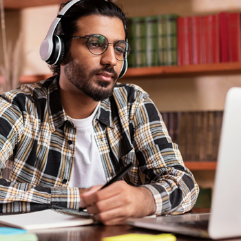 Man wearing glasses and headphones, looking at laptop and taking notes