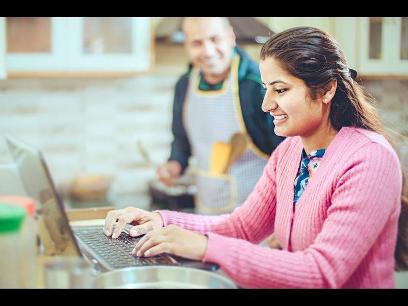 Young woman on laptop in kitchen with man in background