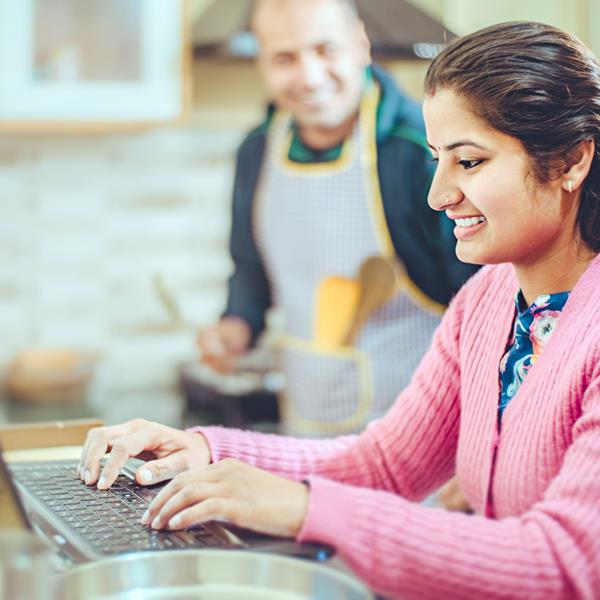 Young woman on laptop in kitchen with man in background