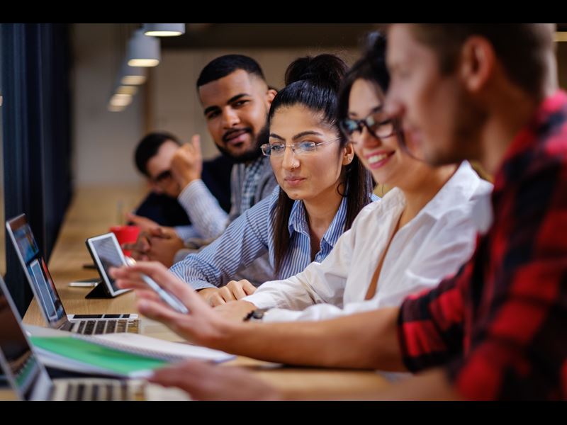 Five people working at desk, looking over at each other's devices