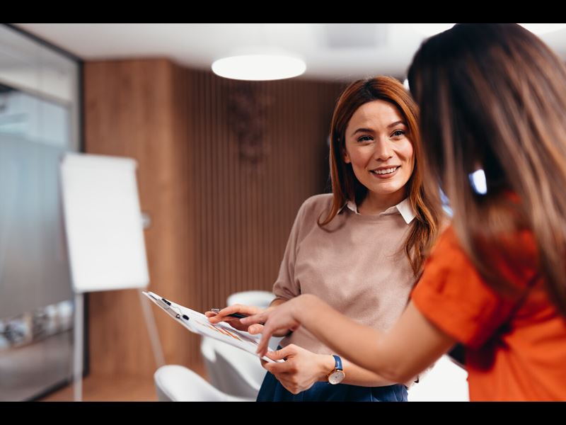Two Female Professionals Smiling And Talking While Holding Clipboard