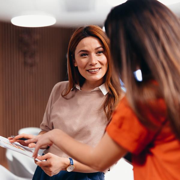Two Female Professionals Smiling And Talking While Holding Clipboard